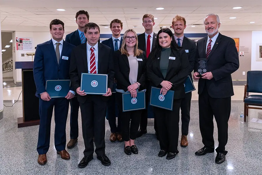 Students in business attire pose holding awards with a government seal on them.