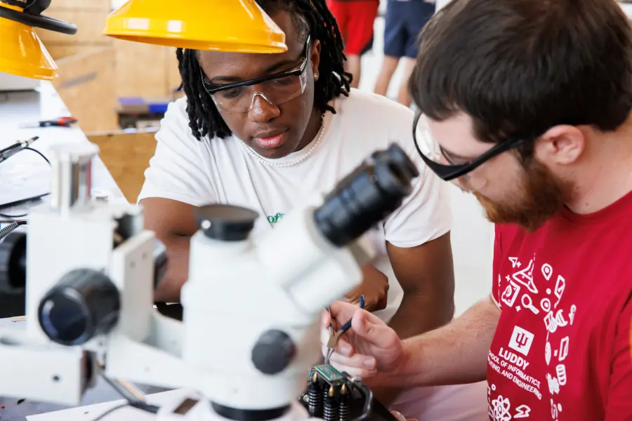 Two students work at a workshop bench filled with microscopes and fabrication equipment.