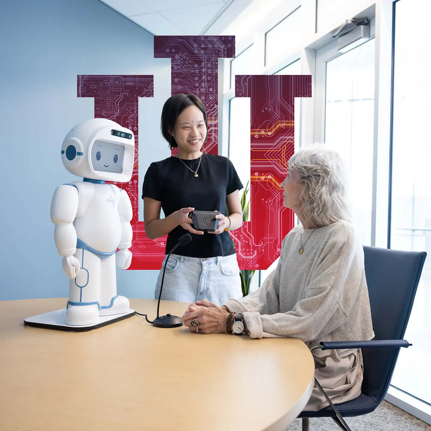 A female student stands next to a table where an older woman sits looking back at her. A white robot stands on the table next to both of the women, the student holds a controller and a microphone sits on the table. A large IU Trident stands behind the student.