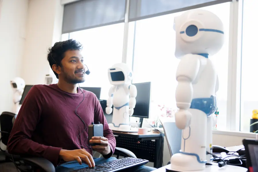 A male student sits with a headset and controller in front of a white robot.