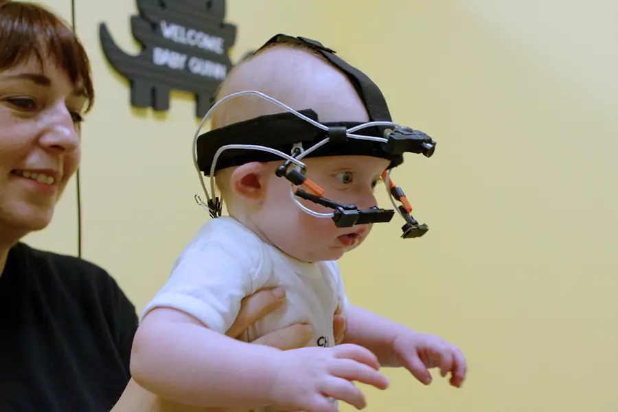 A baby wearing a headband with wires attached looks forward while a woman holds him.