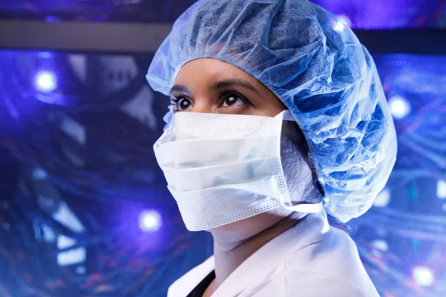 A woman wearing a surgical mask and hair net stands backlit by blue and purple light.