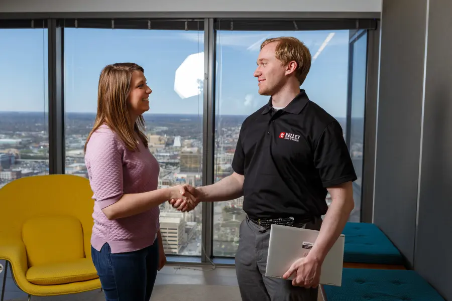 A male student wearing a Kelley School of Business polo shakes a womans hand, the skyline is visible through the window behind them.