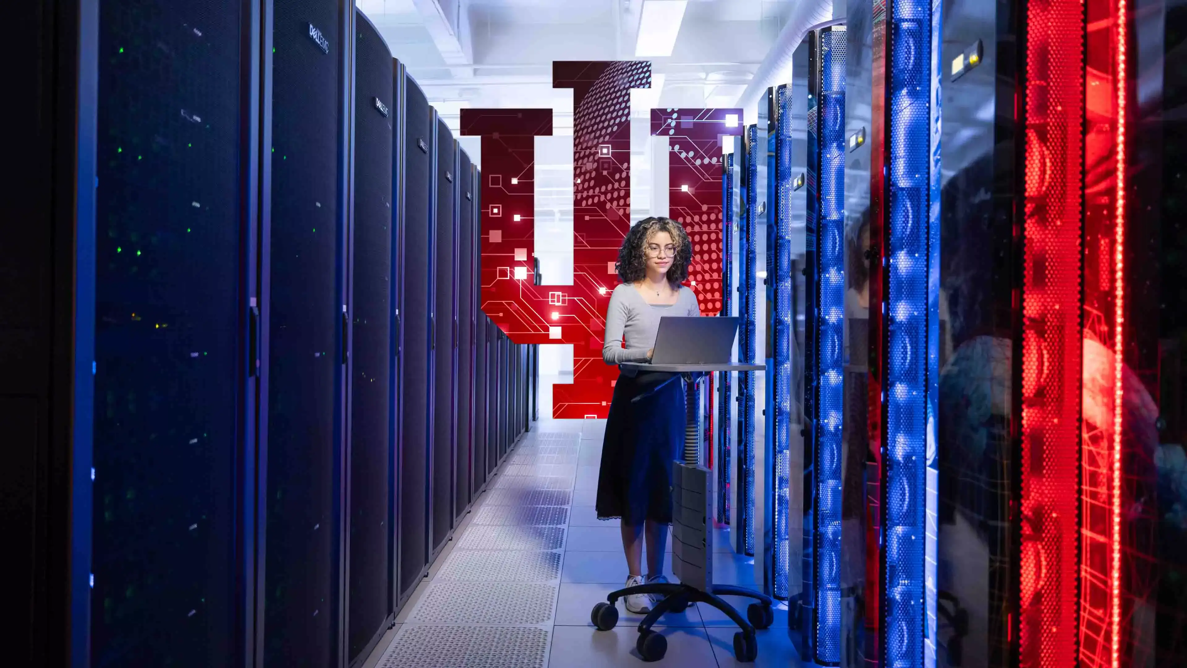 A woman works at a kiosk next to a supercomputer with a large Indiana University trident displayed behind her.