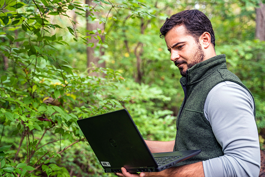 An IU researcher studying the climate, stands in the woods looking at his laptop.