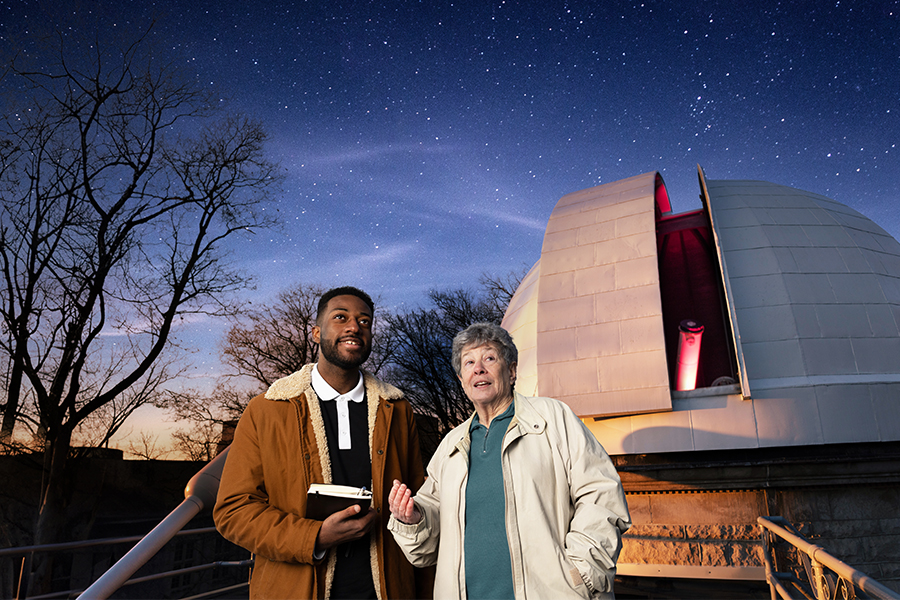 Two people stand looking at the sky in front of the Kirkwood Observatory. A trident is in the background.