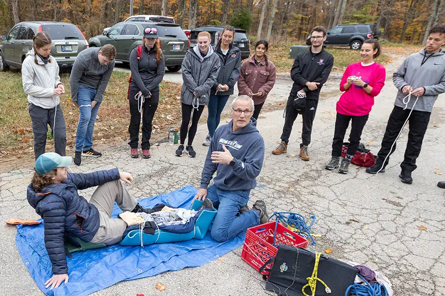 A teacher shows and explains how to splint a mans leg outdoors while a group of students listens.