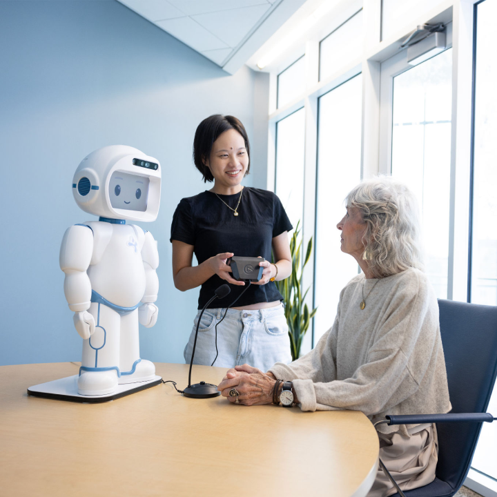 A female student stands next to a table where an older woman sits looking back at her. A white robot stands on the table next to both of the women, the student holds a controller and a microphone sits on the table.