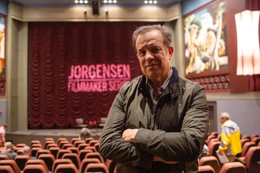 A man with his arms crossed looks back at the camera, an empty theatre stands behind him with a curtained stage.