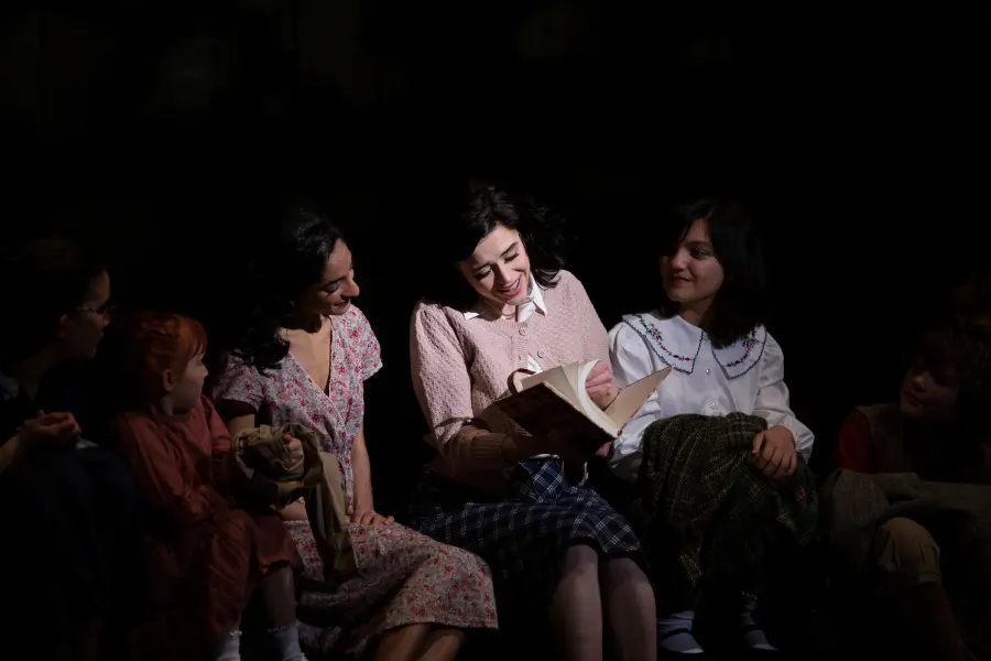 A group of girls appears in the spotlight on a dark stage, they surround a girl as she pages through a book.