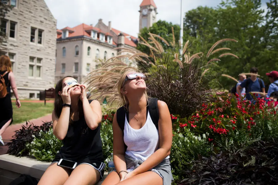 Two girls sit on a limestone wall wearing solar eclipse glasses and peering up at the sky, the bell tower is visible in the distance behind them.