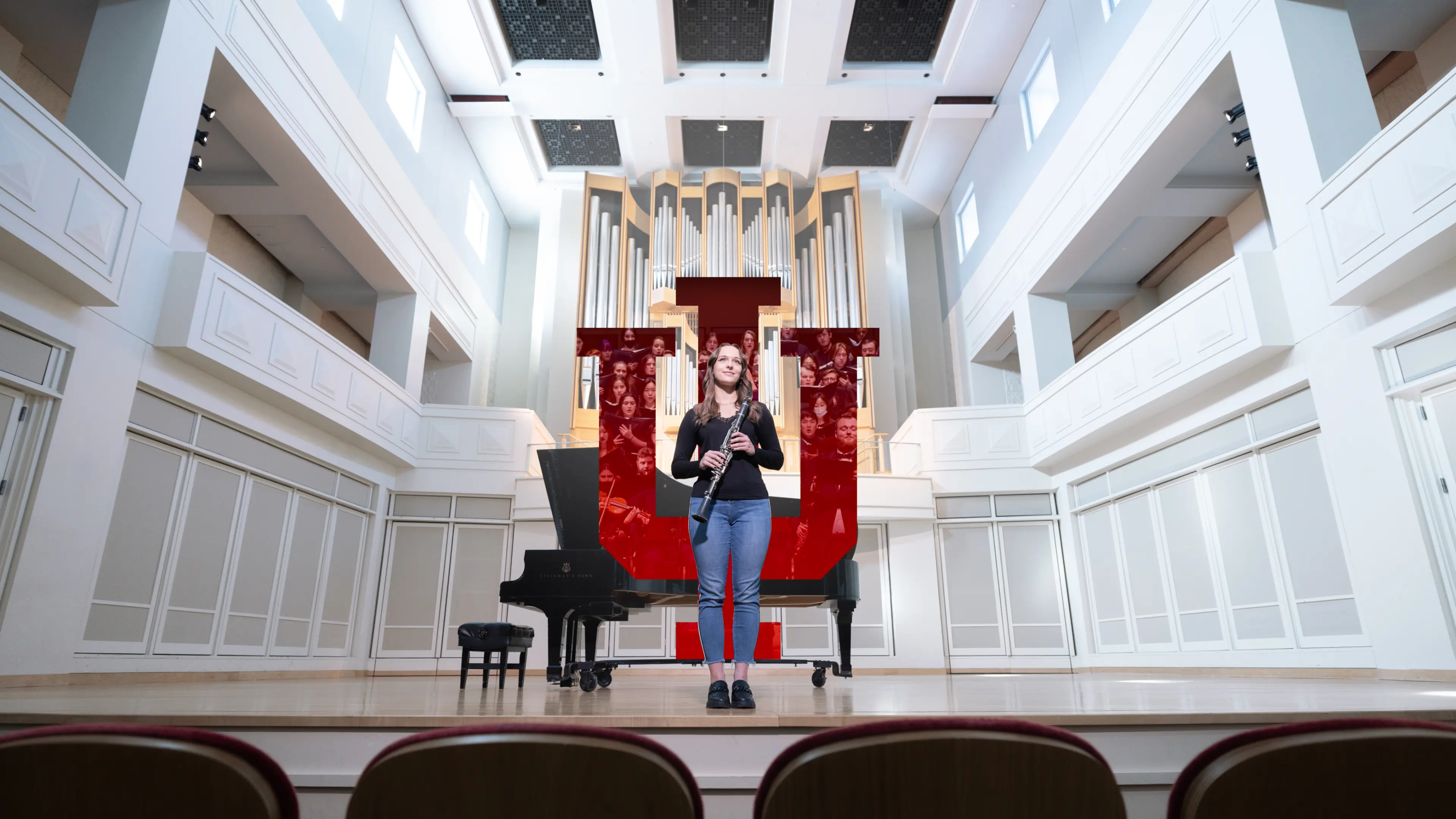 A female student stands centerstage holding a clarinet, she looks out to the performance hall in front of her. Behind her an IU trident shows a full symphony playing.