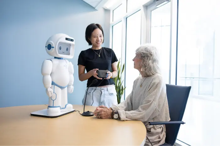 A female student holding a controller stands next to a table where an older woman sits. On the table next to them a white robot stands.