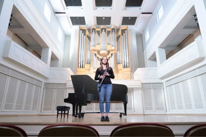 A female student holds her instrument while standing centerstage, she looks out at the performance hall in front of her.