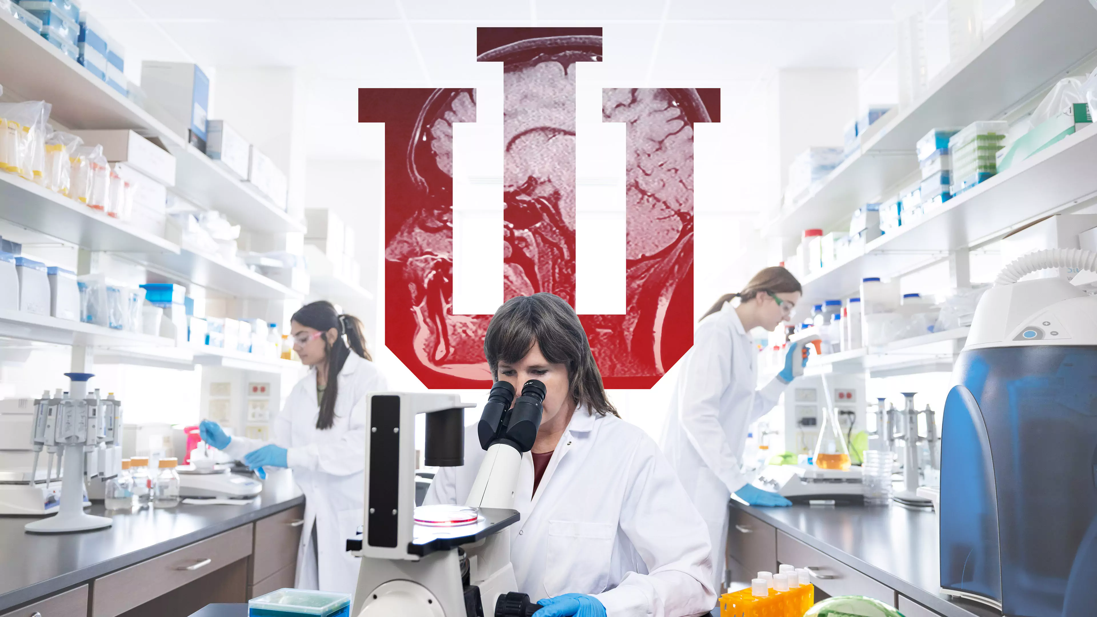 A female researcher wearing a white lab coat peers into a microscope, the lab space behind her is filled with equipment and two other researchers. A large IU Trident stands behind her with a brain scan image within it.