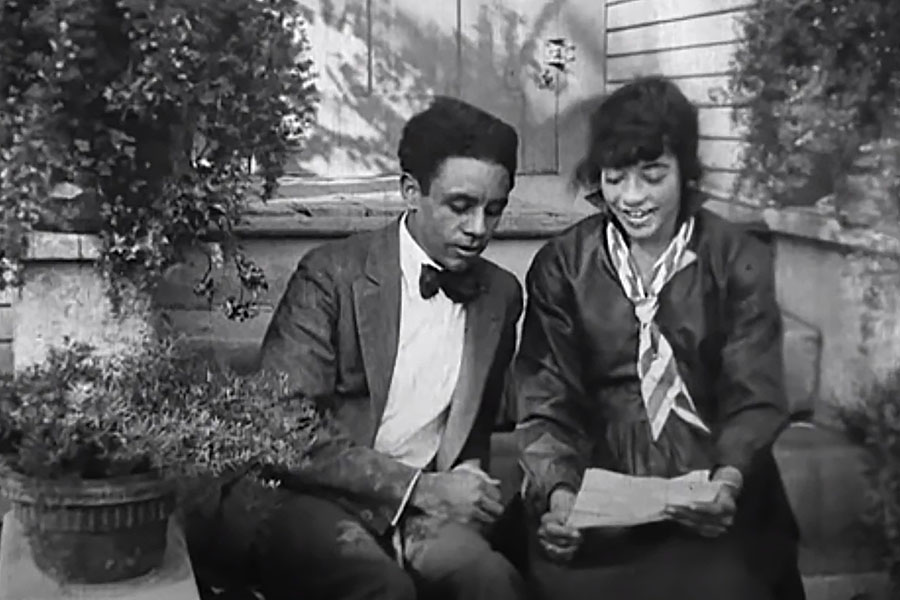A still frame from an early Black film showing a man and woman sitting and talking.