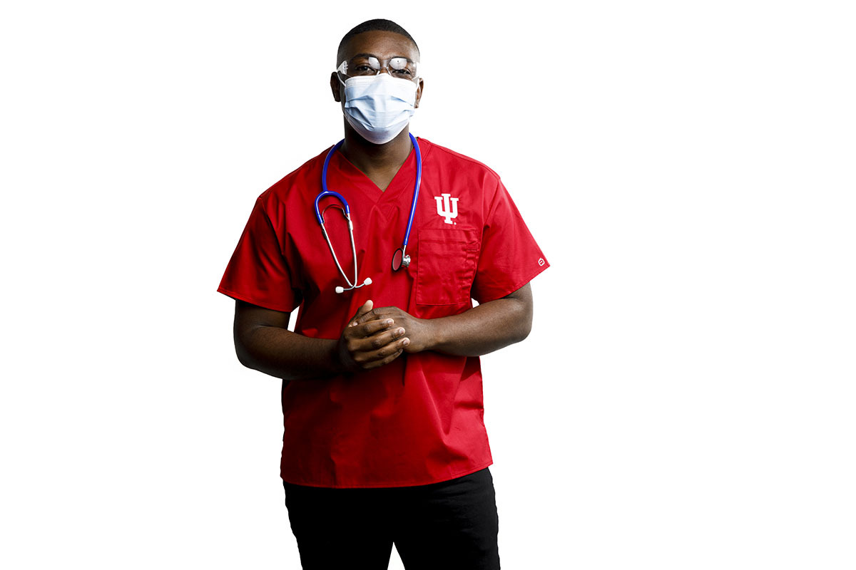 A man faces the camera head-on, with his hands casually clasped in front of him. He wears a red IU Health shirt, protective glasses, face mask, and has a stethoscope draped around his shoulders. 