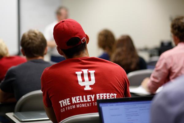 Taken from the back of a classroom, students, including a business student wearing a Kelley Business School shirt that reads The Kelley Effect, attentively look towards the professor teaching. 