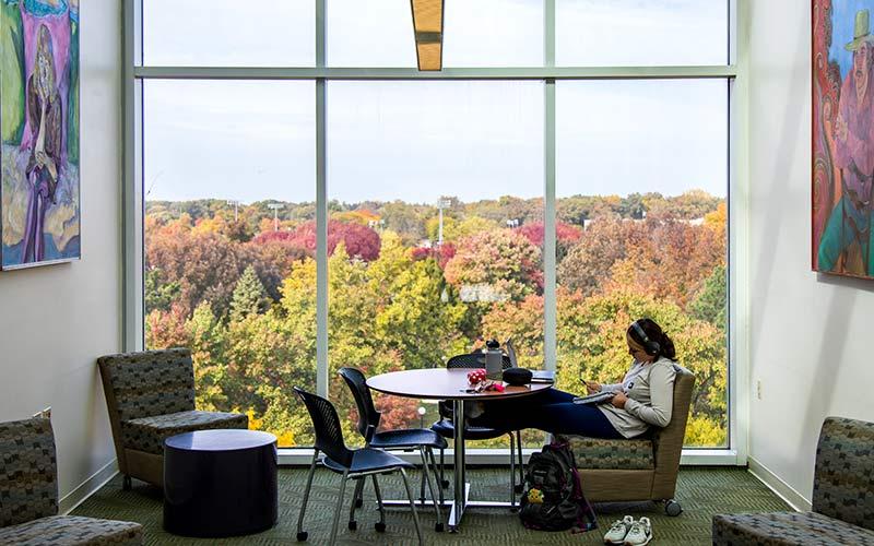 A student sits studying with headphones in front of window overlooking colorful fall trees.