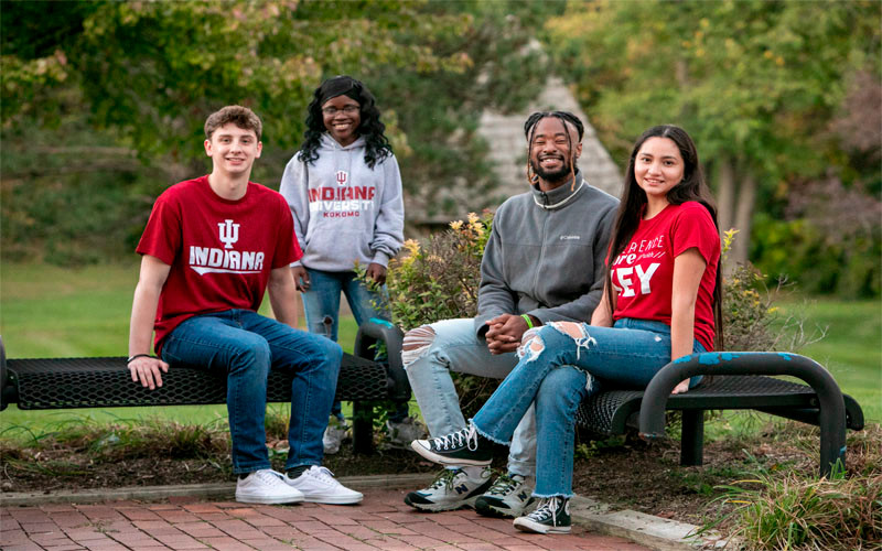 Three students sit on outdoor benches and one student is standing. Three of them wear IU shirts.