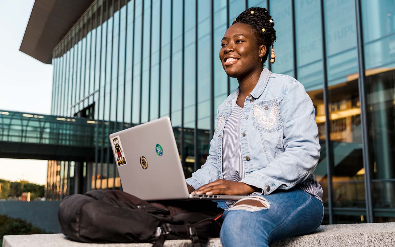 A female student sits on a limestone wall outside of the IU Indianapolis campus center, she smiles into the distance while typing on her laptop.