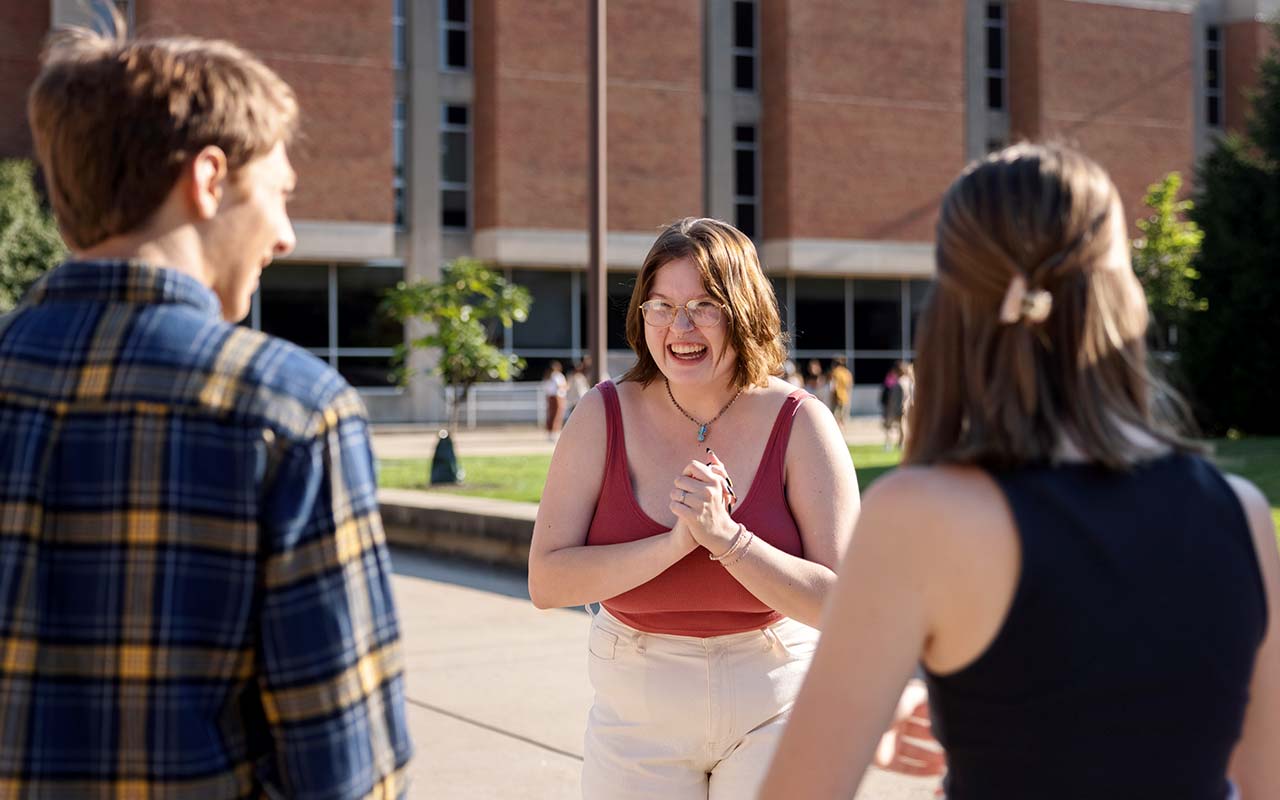 A female student faces the camera and two other students, she laughs, clasping her hands in front of her chest. The IU Indianapolis campus and Taylor courtyard is behind them.