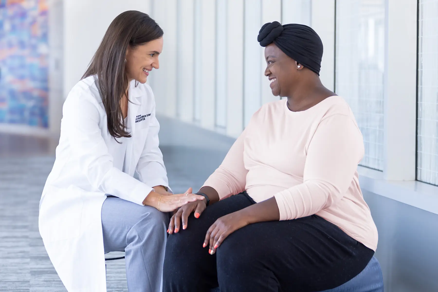 A white female oncologist sits with a black female cancer patient in a bright hallway.