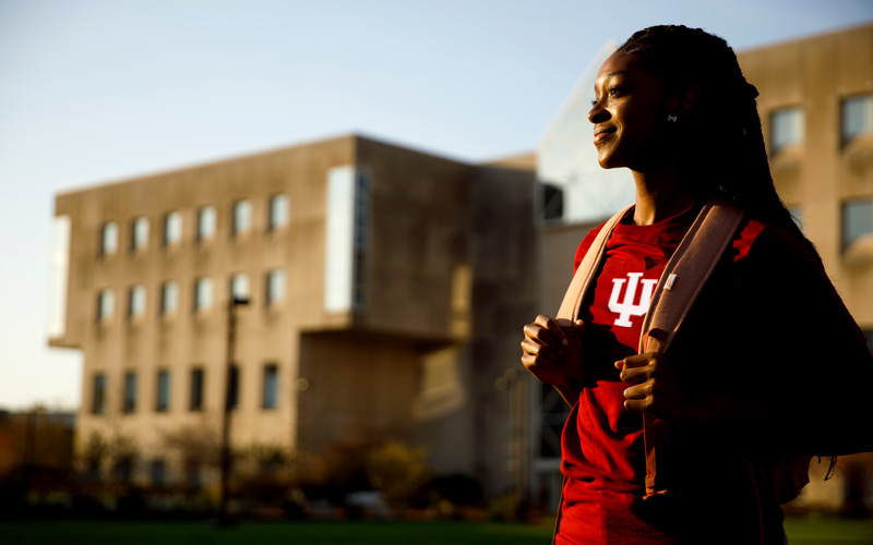 A female student smiles into the distance while standing in front of the University Library at IUPUI, she wears a red shirt with a white trident centered between the straps of her backpack.