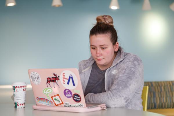 A female student sits alone at a table in a group area, looking at her laptop with a serious face.