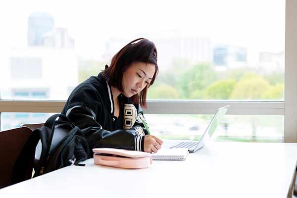 A female student sits in the IU Indianapolis University Library, she is focused on writing in her notebook. The city skyline is visible in the background.