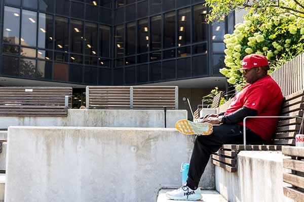 A male student sits at a bench while typing on his laptop.