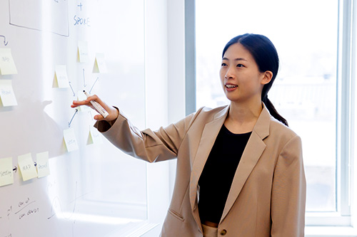 A female student stands pointing at post it notes placed on a whiteboard.