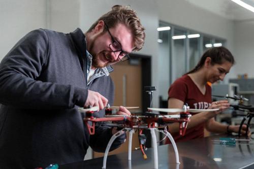 Two students smile as they tinker with drones on a workbench.