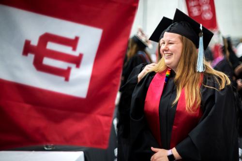 A graduate smiles wearing her cap and gown near a crimson flag with the IU trident proudly displayed.