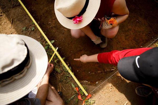 Three archeology students looking into and measuring a hole in the ground.