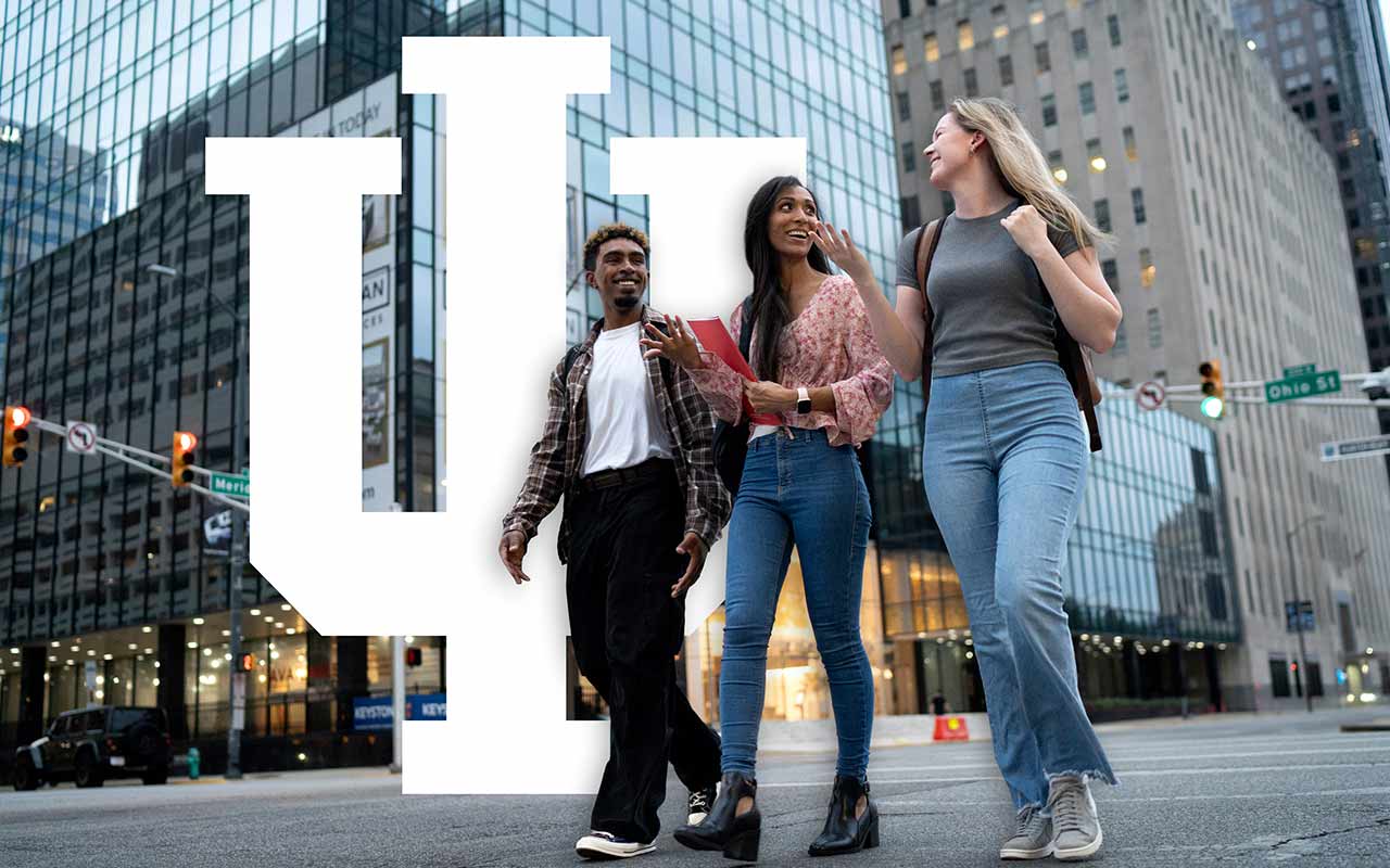 Three smiling students walk through a downtown Indianapolis street with glass buildings and street lights behind them.