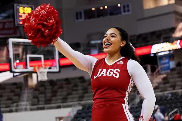 A female cheerleader stands on the basketball court mid-cheer, her crimson uniform has the JAGS wordmark on the chest.