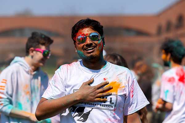 A male student stands, smiling and facing the camera, his hand is on his chest and he's covered in colorful chaulk celebrating the festival of Holi.