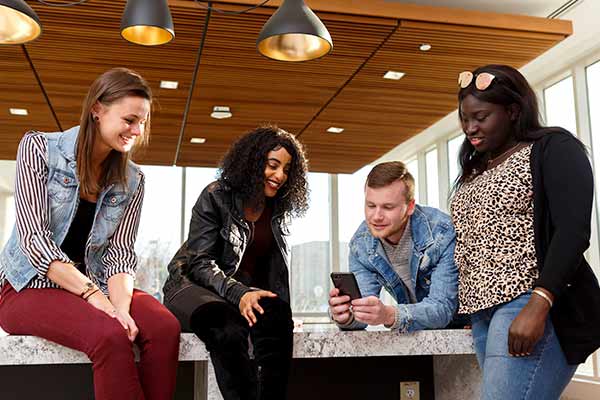 Four students casually gather in a common space of an IU Indianapolis residence hall, one student holds his cell phone for the others to look at.