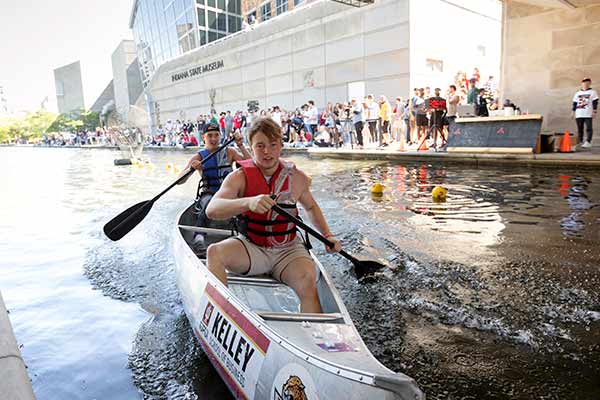 Two students paddle in a canoe during IU Indianapolis' Regatta, spectators line the Indianapolis Canal in front of the State Museum in the background.