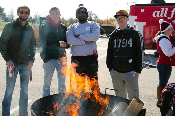 Group of male students standing in front of a fire at a tailgate.