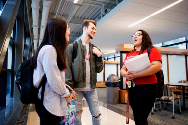 Three students smile and chat while standing in a campus building.