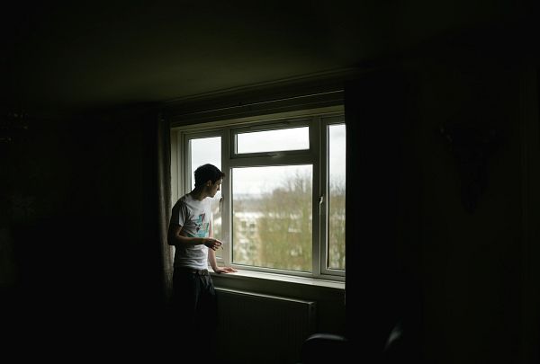 A young man, possibly a student, gazes out a window overlooking campus while holding a lit cigarette.