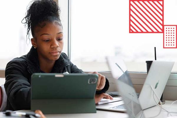 A student sits at a desk viewing information on a laptop and tablet.