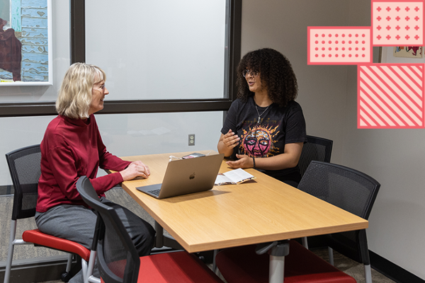 Two adult women having a discussion at a table. An open laptop is between them along with some papers.