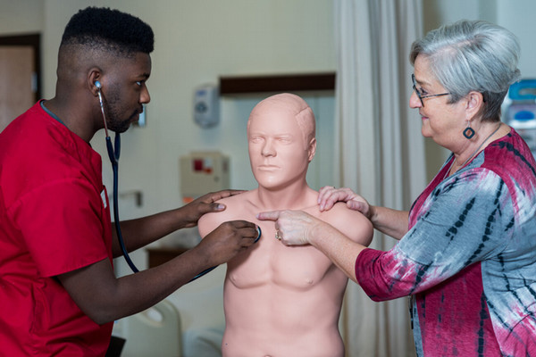 A student holds a stethoscope to a dummy's chest while a teacher explains.